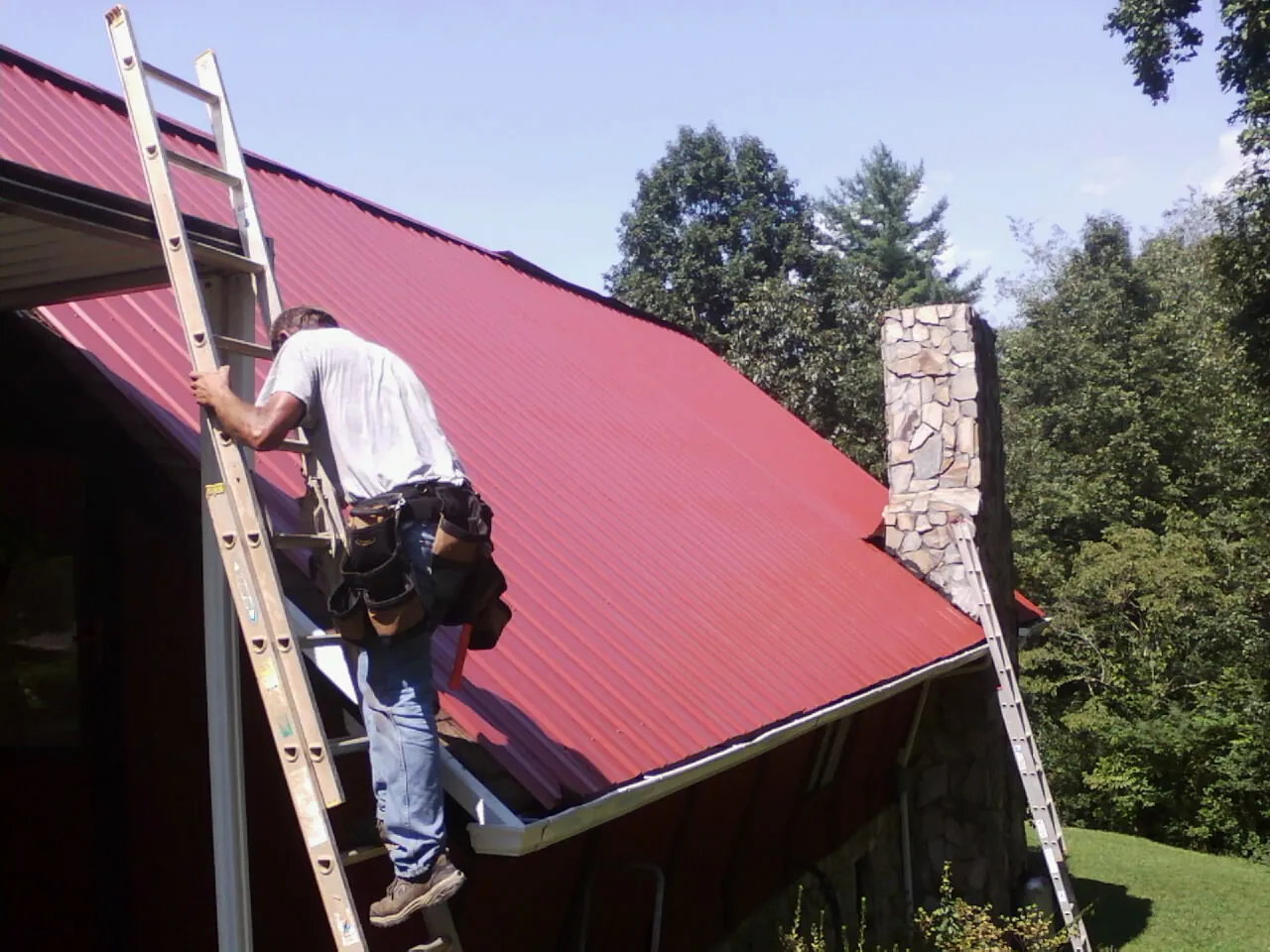 Roofer on a ladder beside a red metal roof and stone chimney — Soffit & Fascia Repair by Roofing Brothers in Ethridge